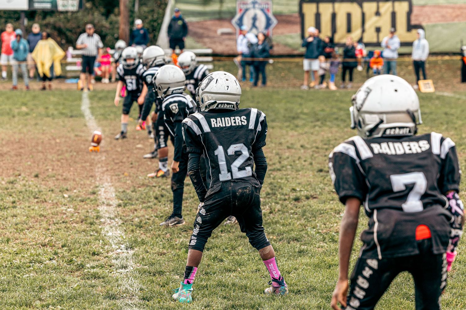 Youth football players lined up in formation wearing dark uniforms with pink cleats.