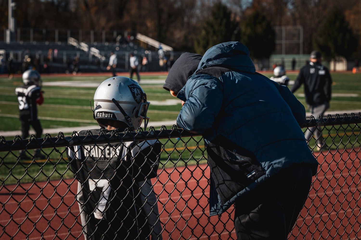 A row of football players watches the game from behind a red fence on the sidelines.