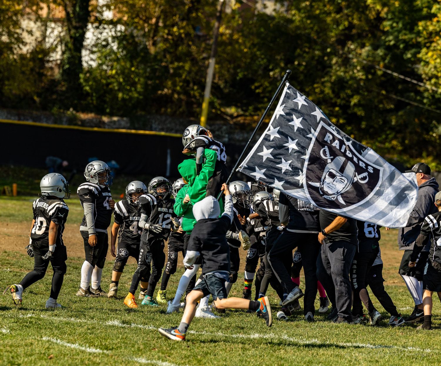 Players celebrate on the sideline while someone waves a Raiders flag on a sunny football field.