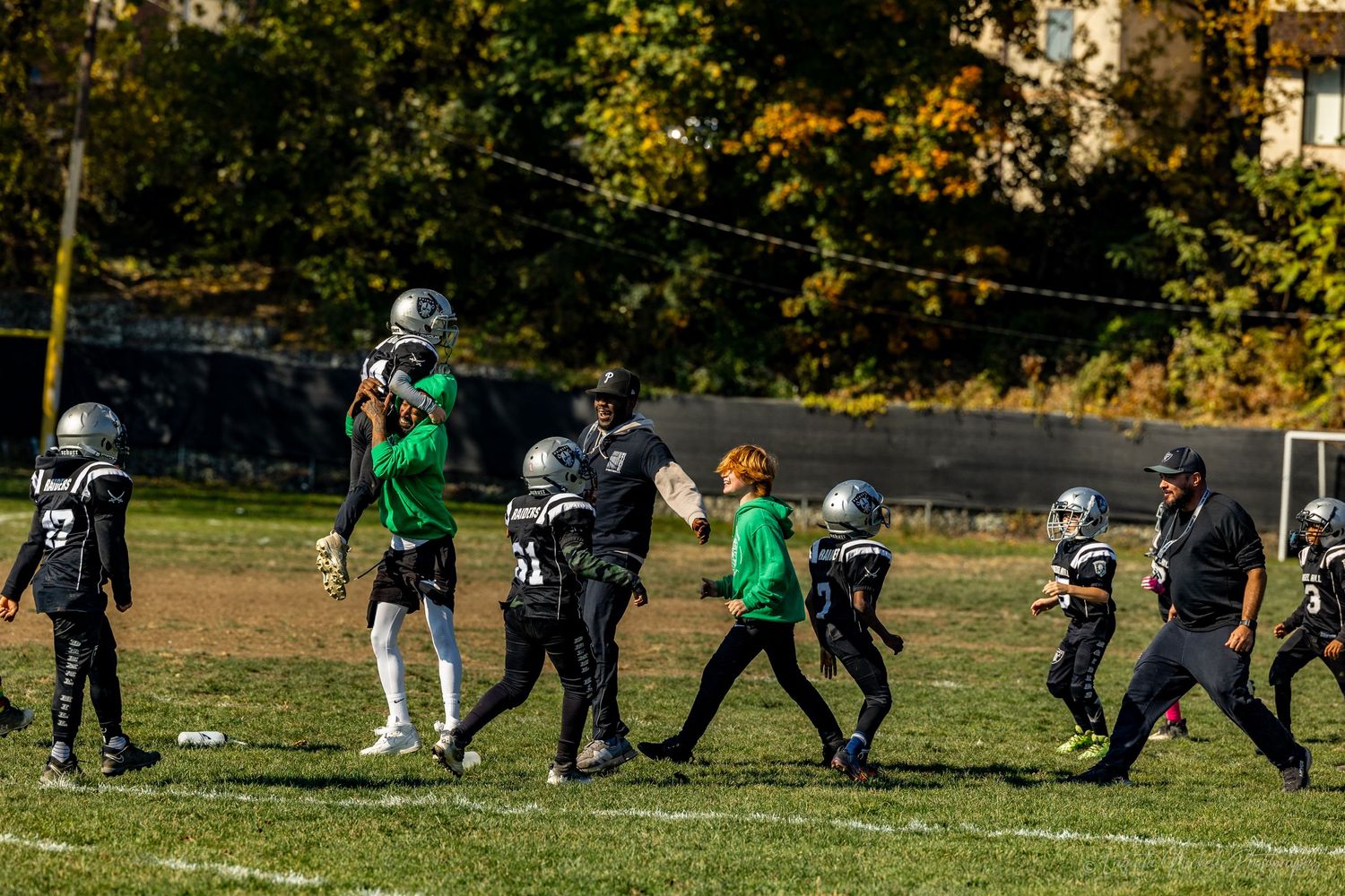 Football players and coaches walk across a grass field during practice on a sunny autumn day.