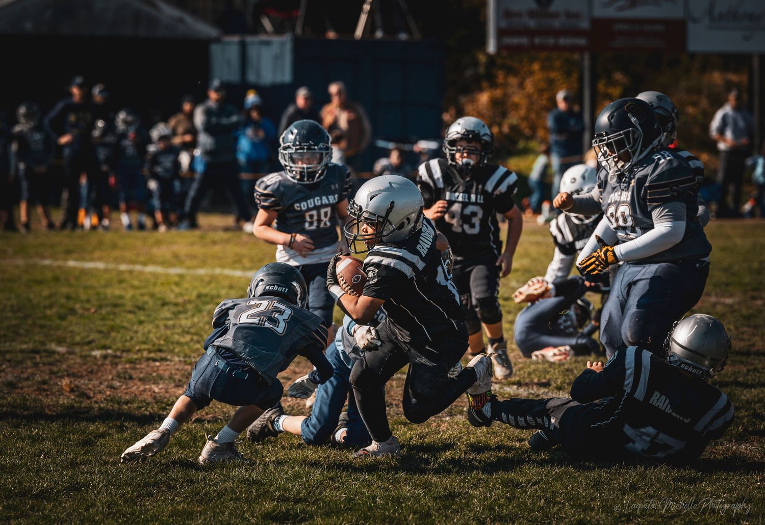 Youth football players engage in a tackle during an intense game on a sunlit field.
