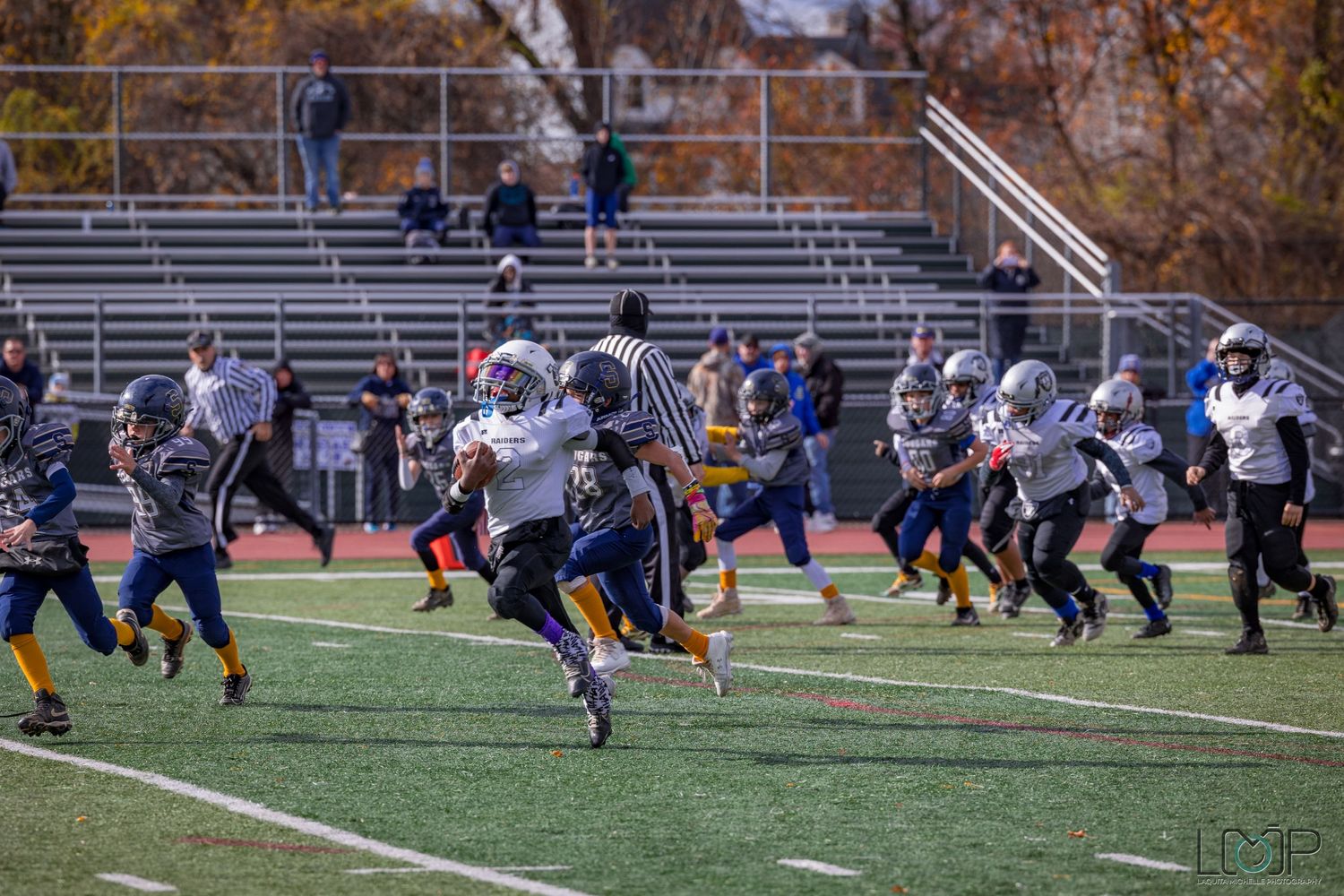 Football players lined up in formation during an autumn game with stadium bleachers visible.