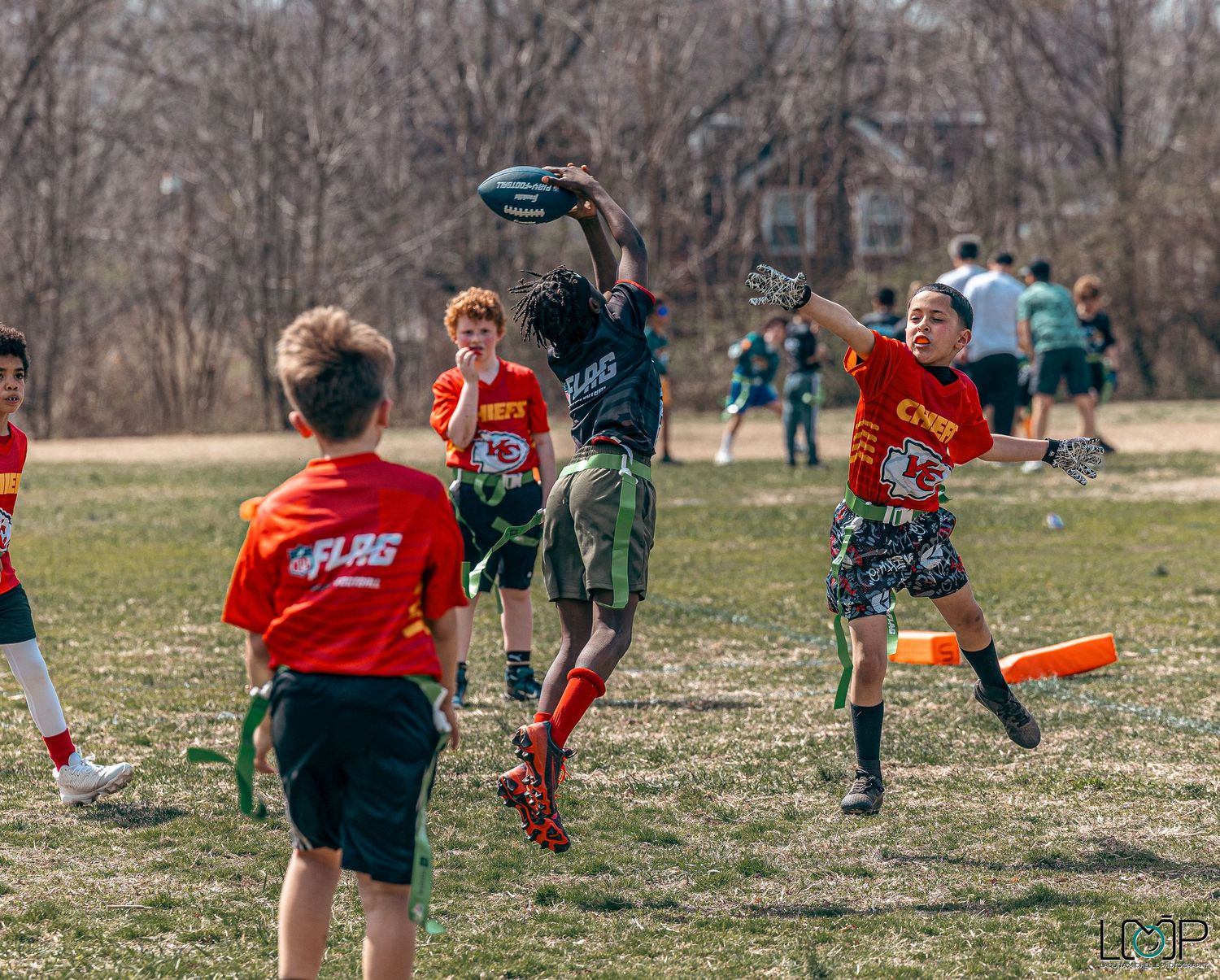 People in red and black jerseys playing an outdoor soccer match on a grass field in early spring.