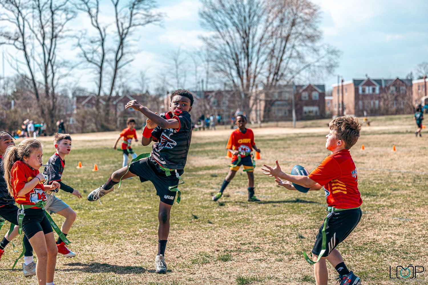 Group playing flag football outdoors on a grassy field during a sunny day with bare winter trees in background.