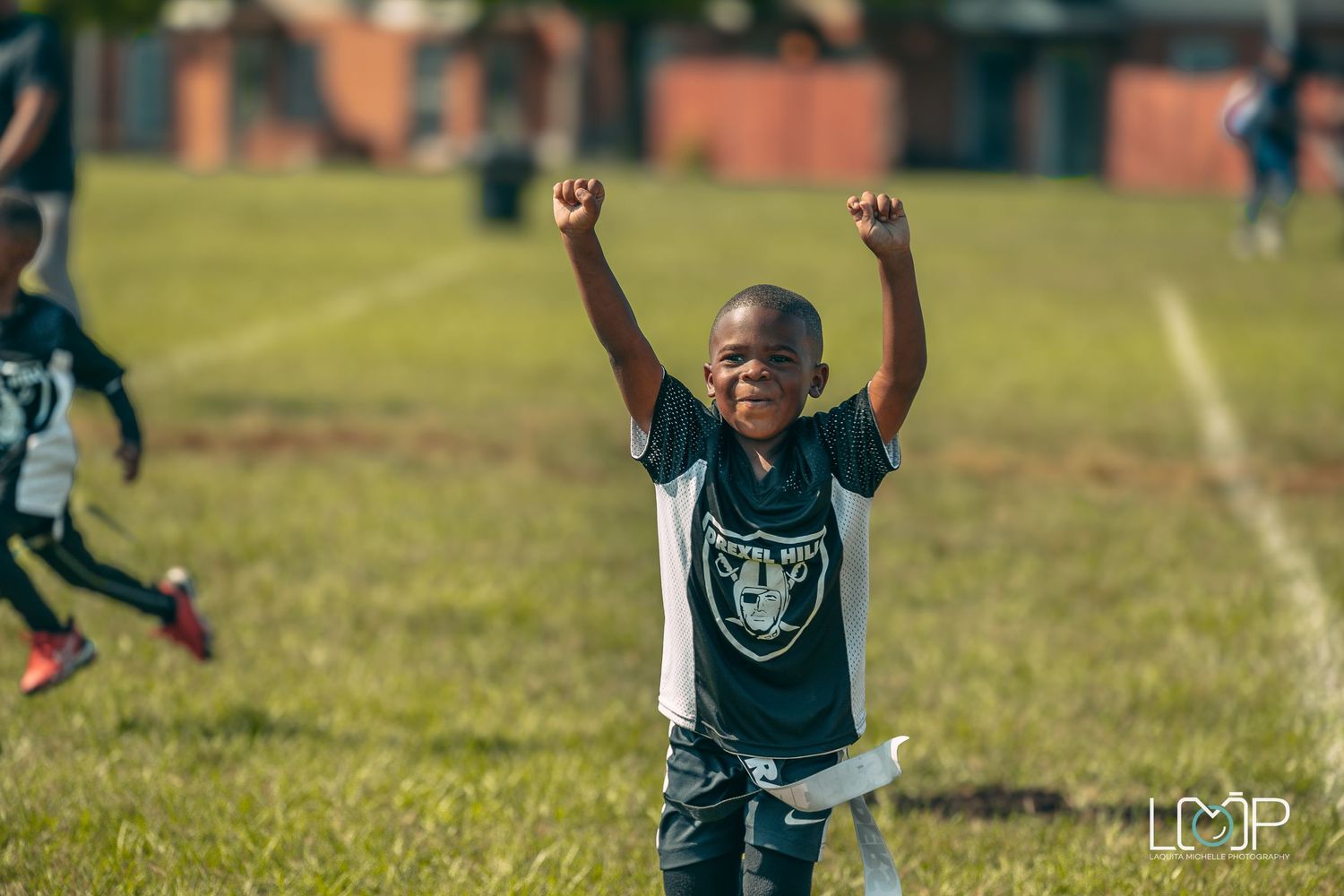 Young player celebrates with raised arms wearing Raiders-style jersey on football field.