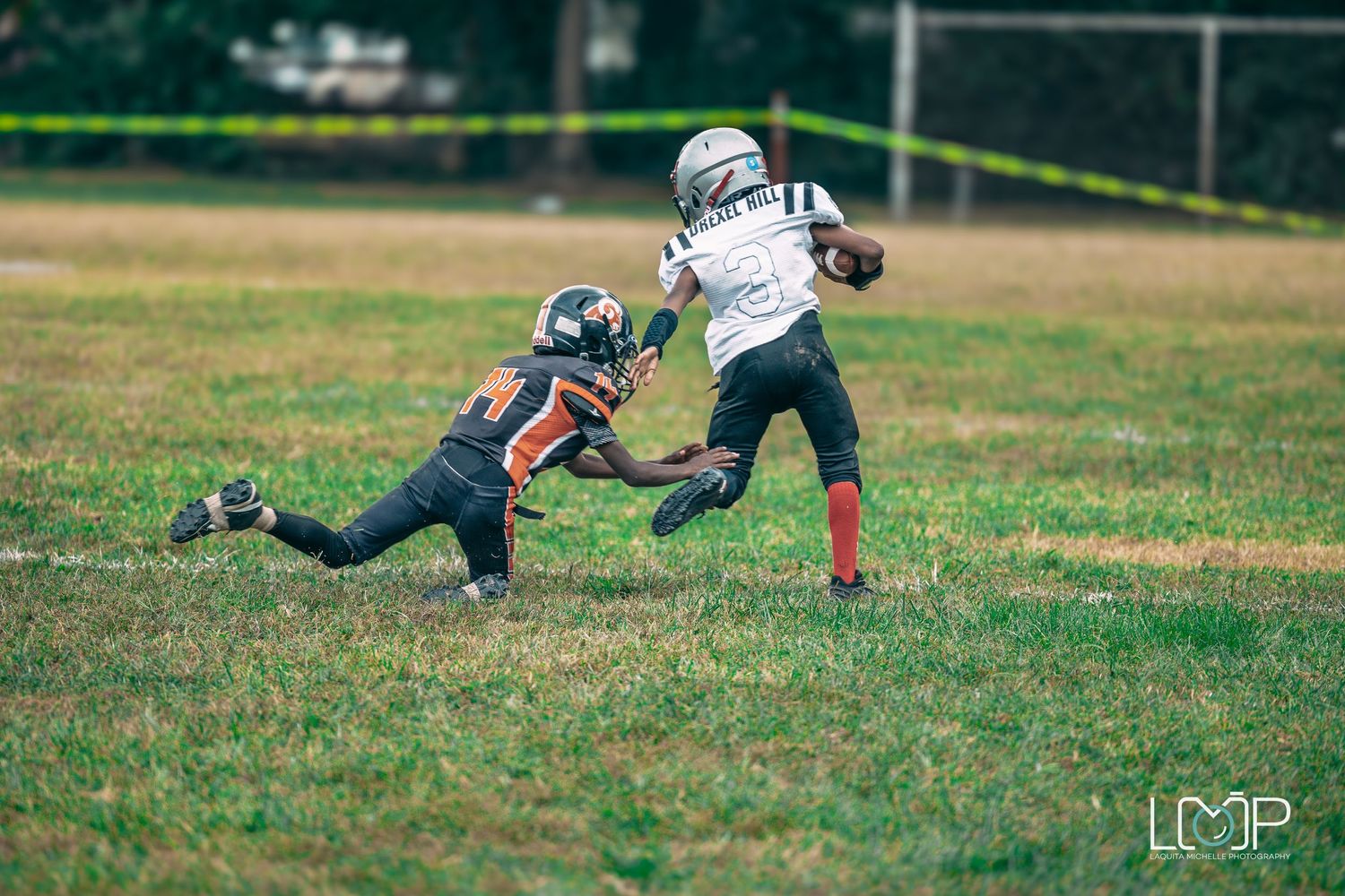 Youth football players in action with one attempting a diving tackle on the grass field.