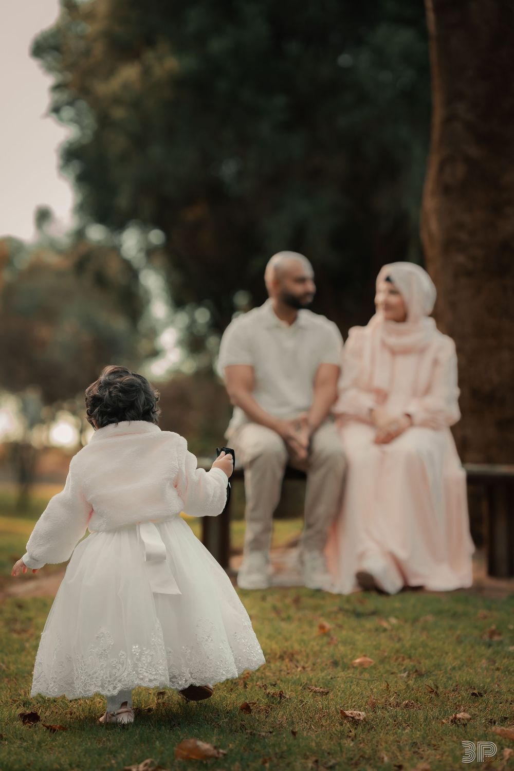 A toddler in a white dress walks toward her parents sitting on a bench in a park during a family photo session.