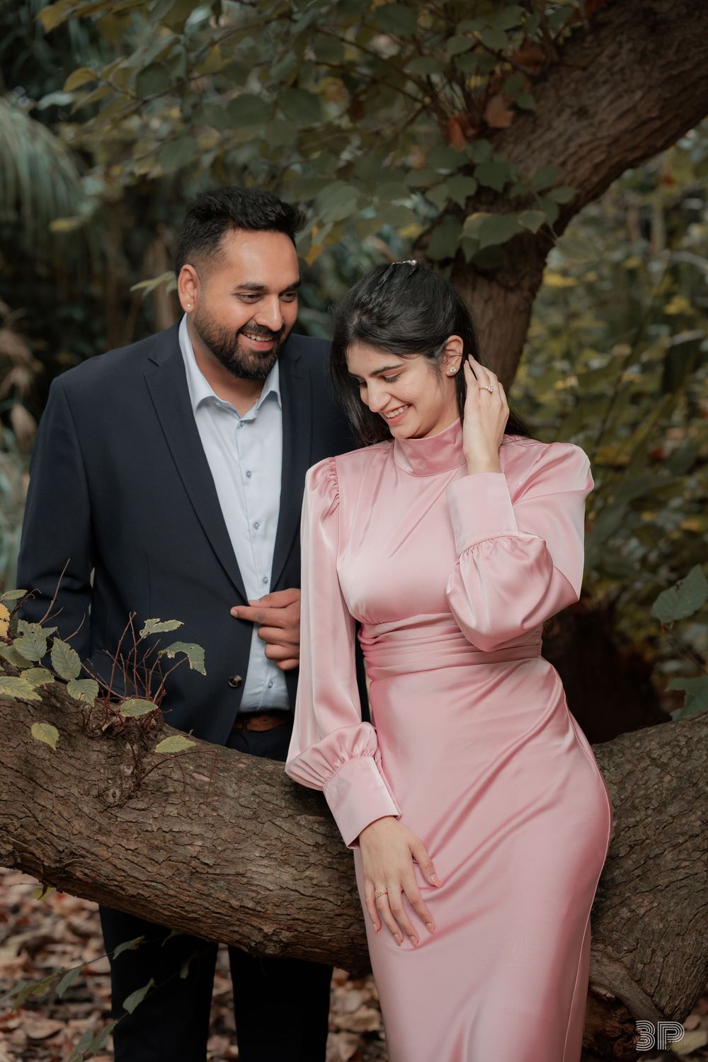 A couple poses near a tree, man in dark suit, woman in elegant pink satin dress, sharing a romantic moment outdoors.