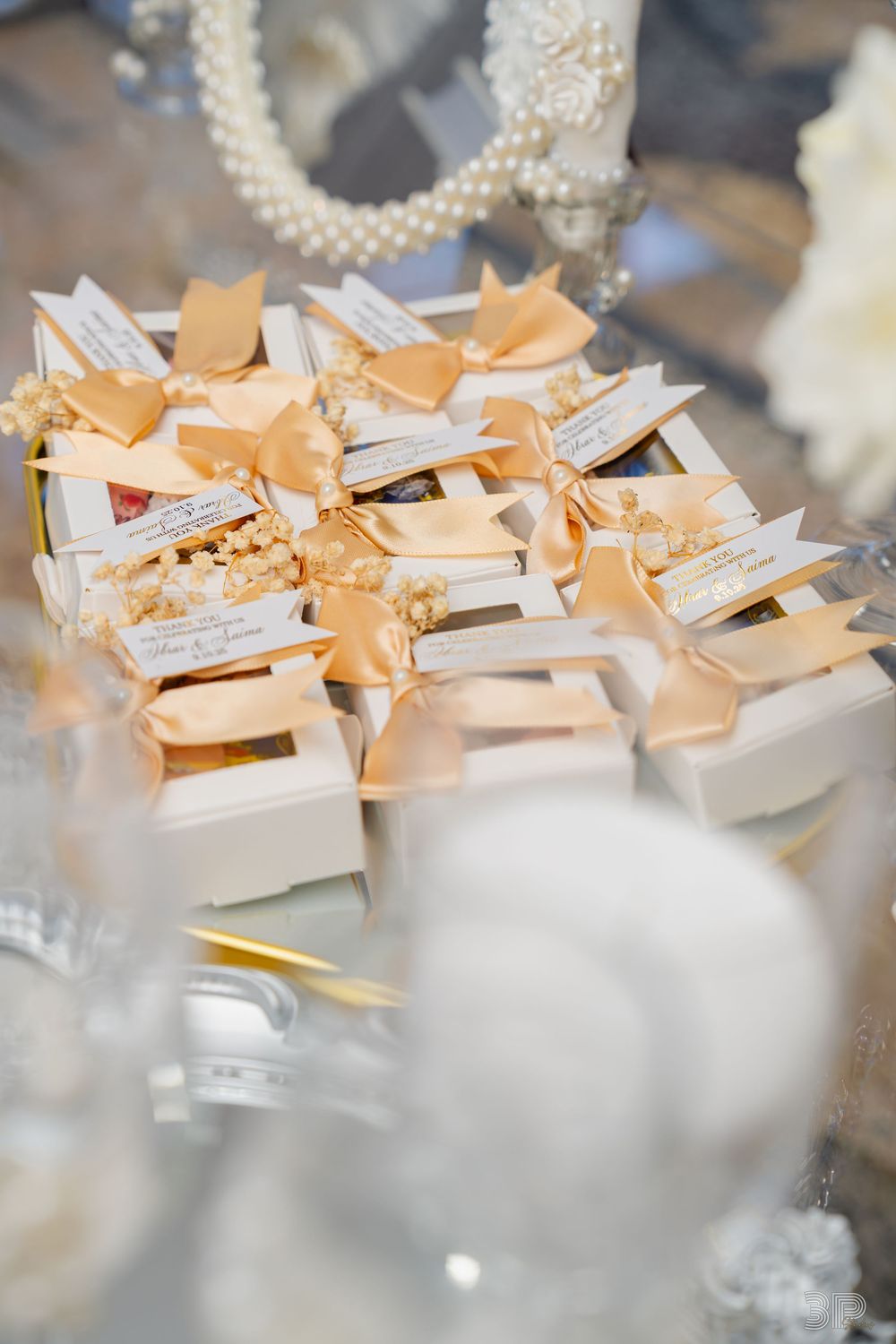 Elegant white wedding favor boxes with gold satin ribbons and pearl accents arranged on a mirrored surface.