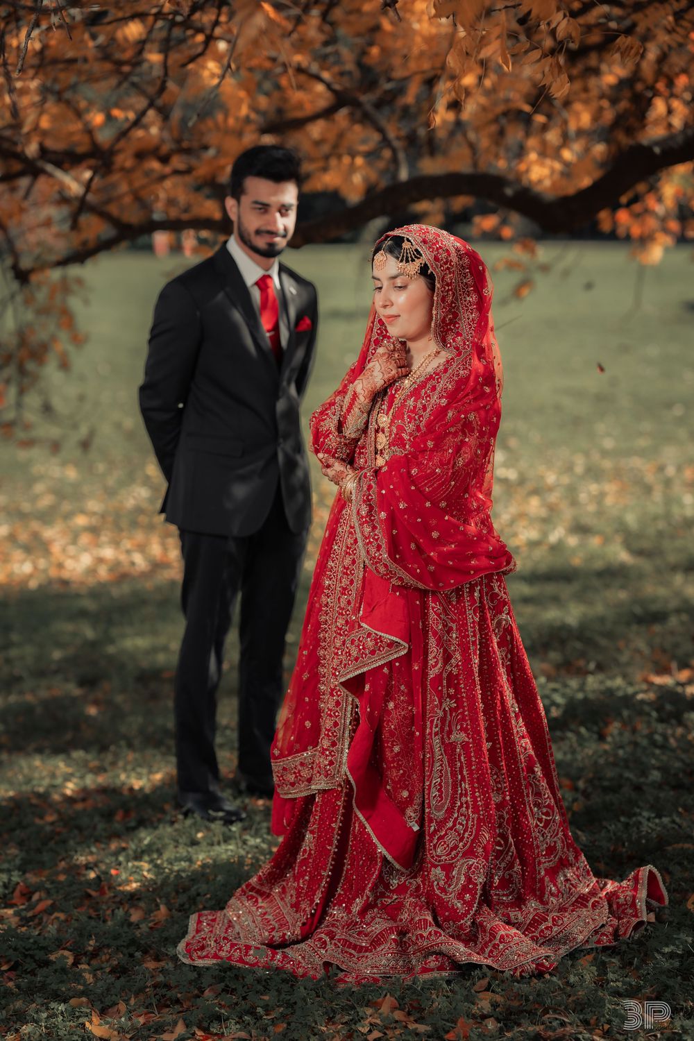 Bride in red bridal lehenga with groom in black suit posing outdoors among autumn foliage.