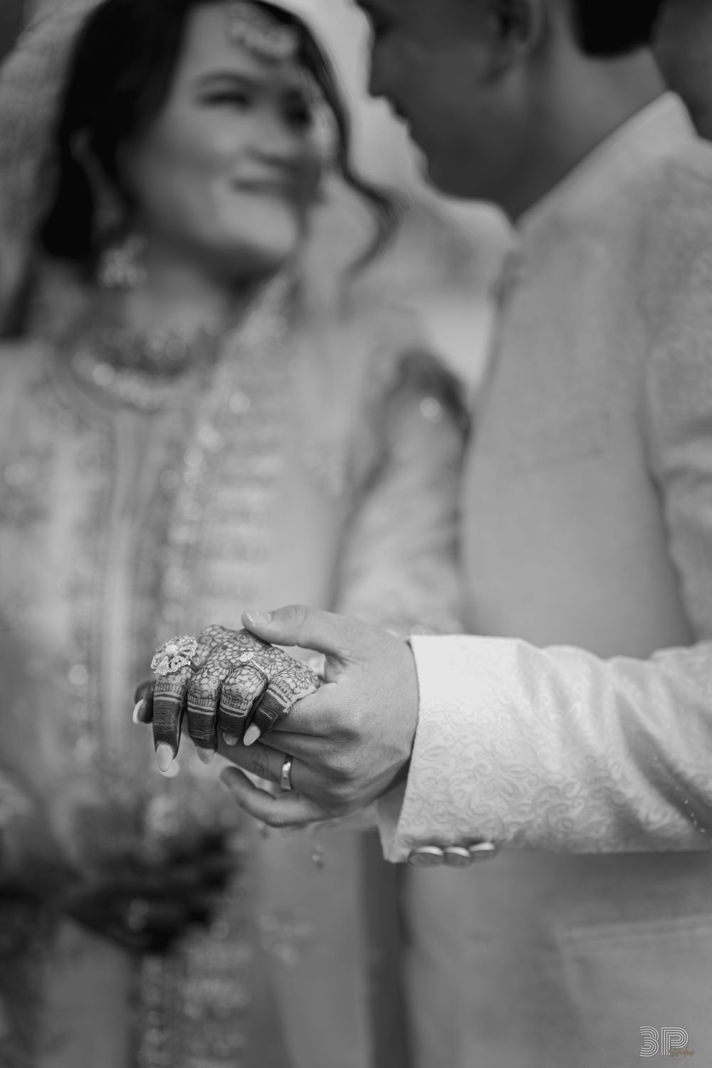Black and white wedding photo of bride and groom holding hands, showcasing intricate mehndi and diamond rings.