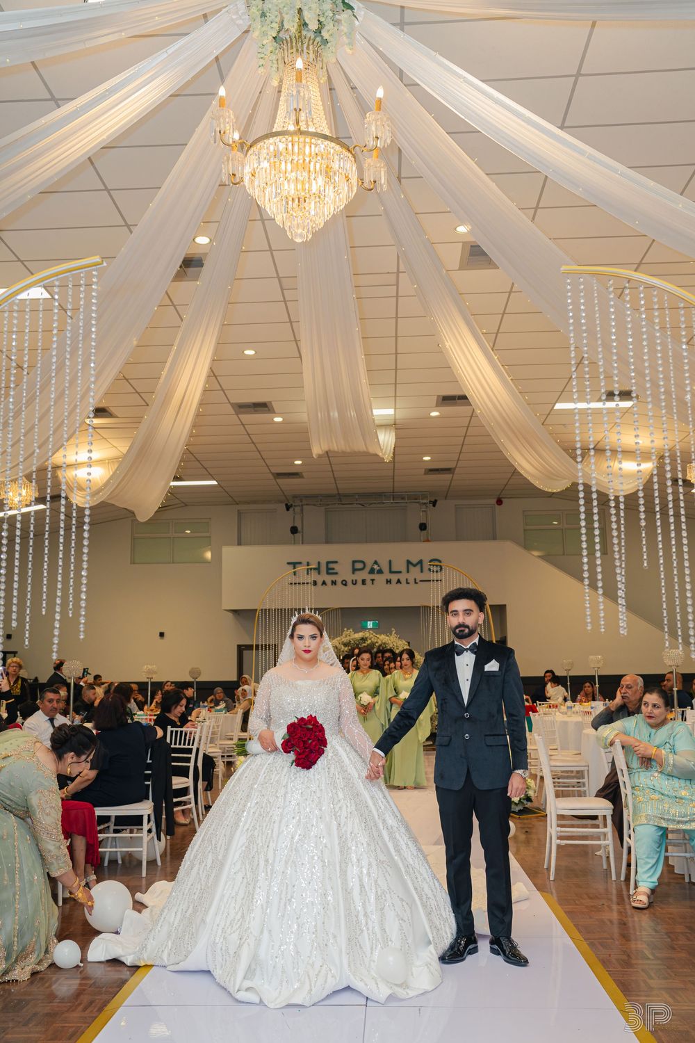 Bride in white ball gown and groom in dark suit walk down aisle at The Palms Banquet Hall wedding reception.