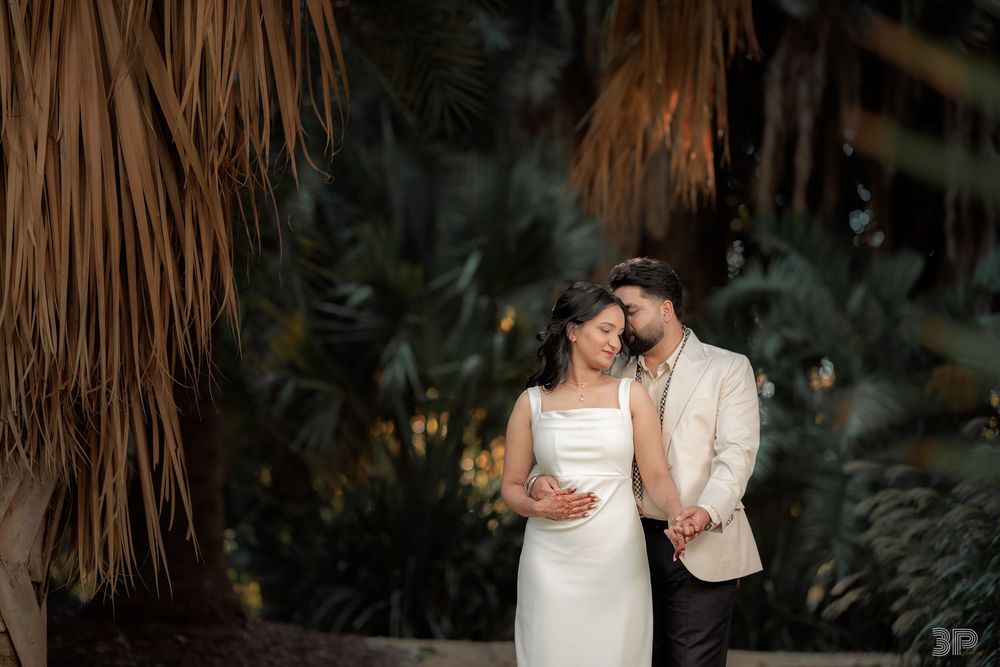 Couple in elegant wedding attire embracing among lush tropical palms, bride in white gown, groom in beige blazer.