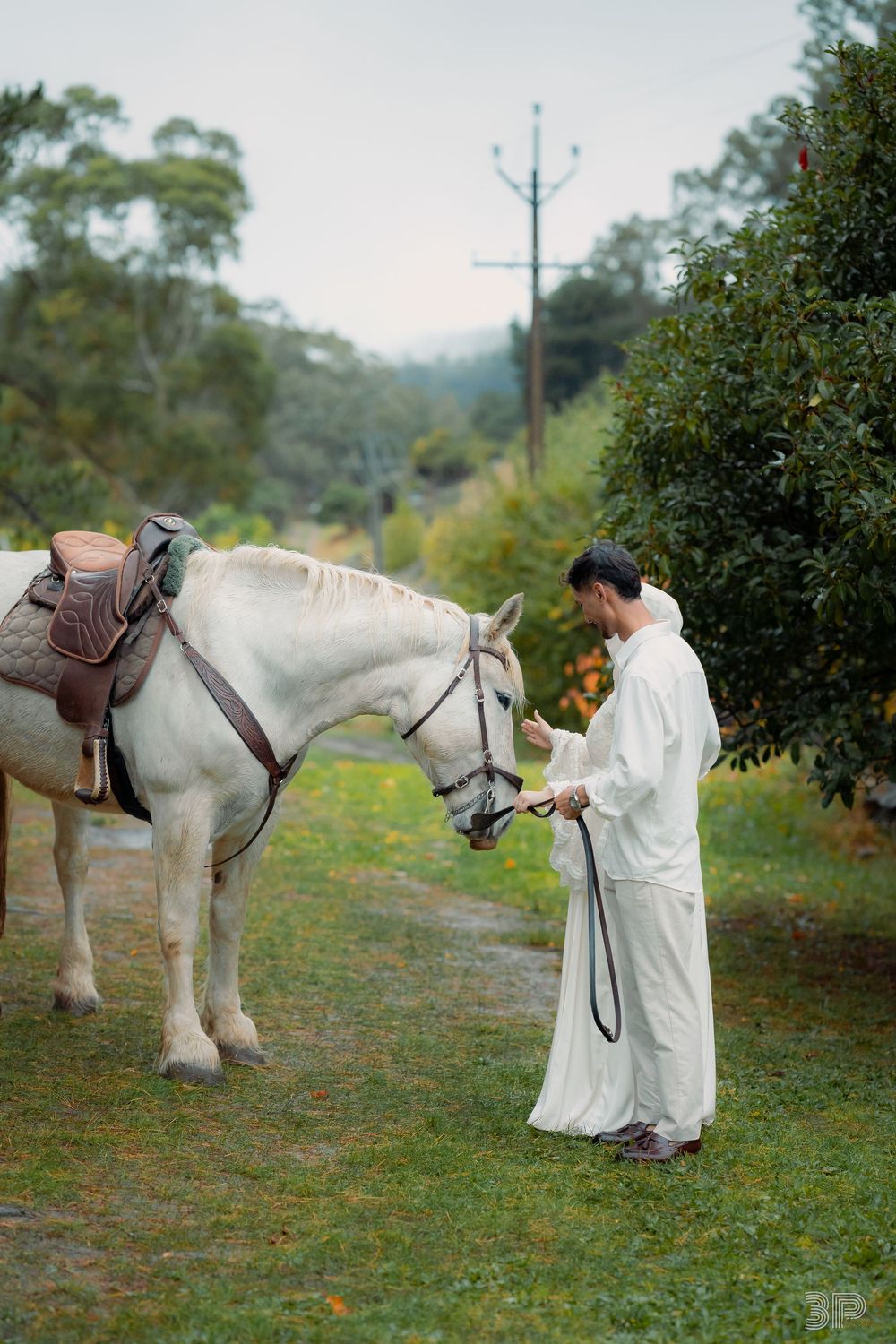Man in white robe holding reins of a saddled white horse in a lush green outdoor setting.