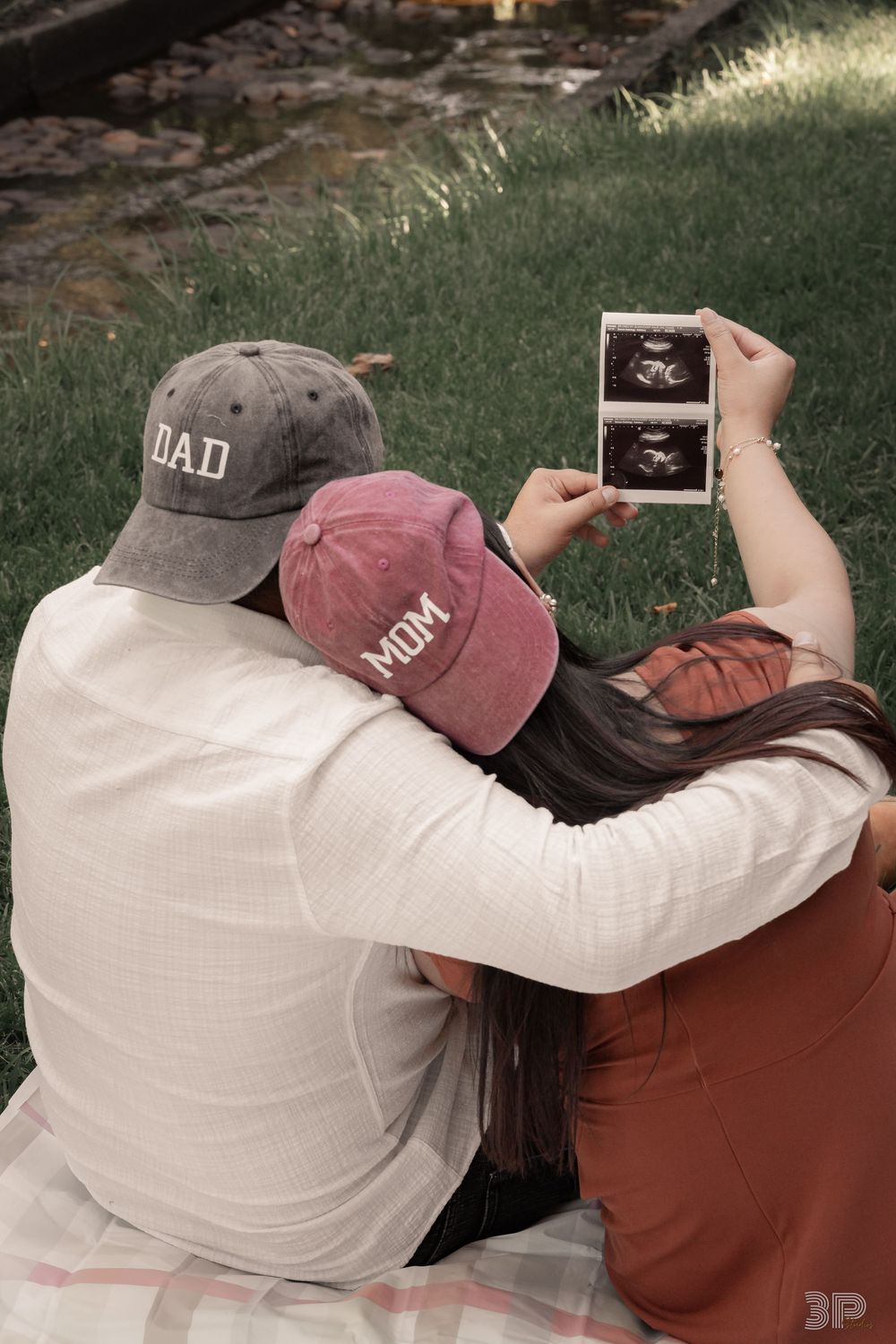 Couple sitting on grass wearing Dad and Mom hats holding ultrasound photos, announcing pregnancy outdoors.