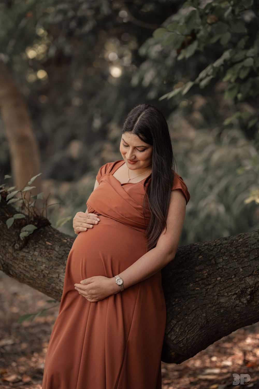 Pregnant woman in rust dress cradling her baby bump, leaning against a tree in a lush green forest setting.