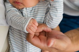A baby's tiny hand grasps an adult's finger while wearing a gray and white striped sleeve.