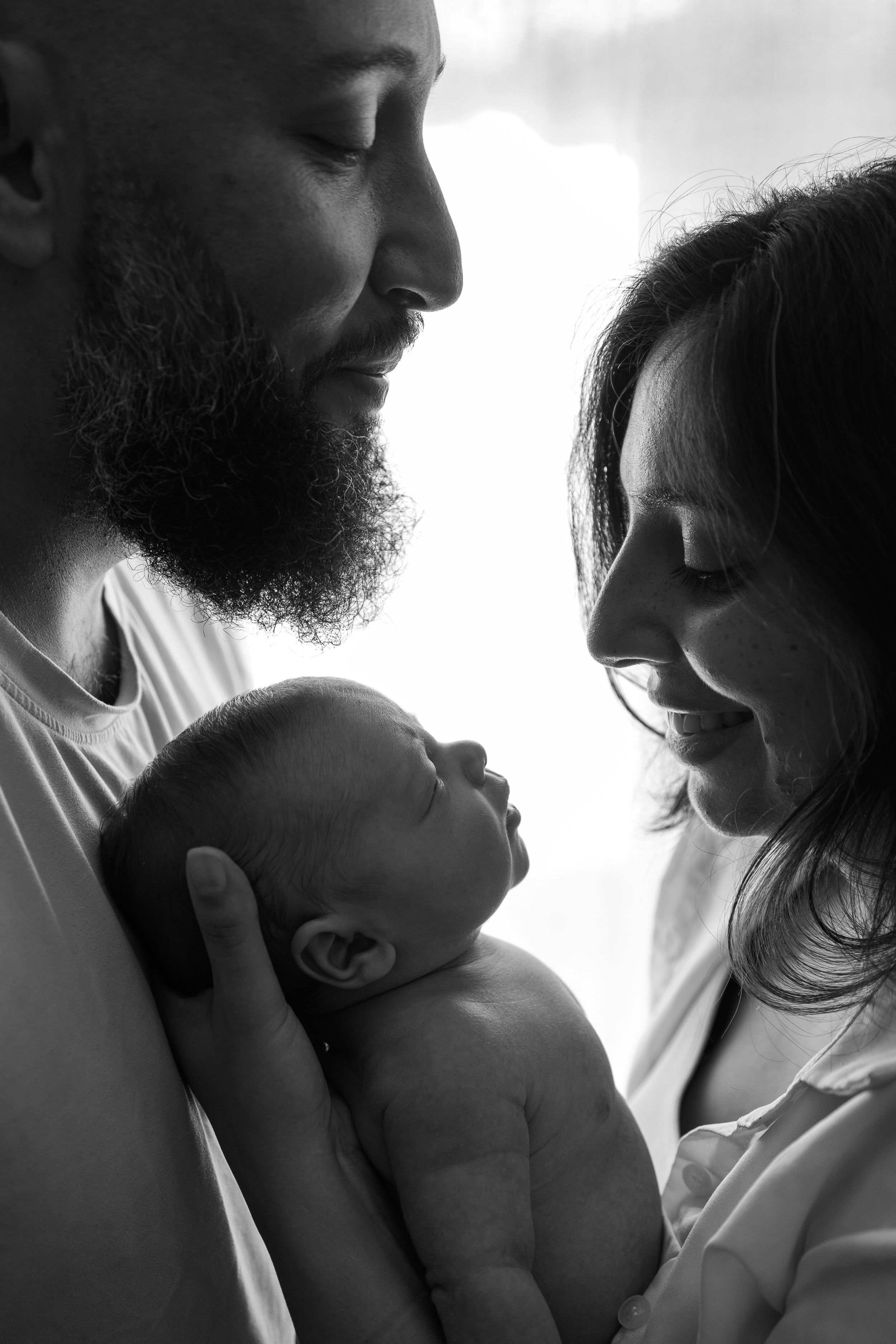Silhouette of parents sharing tender moments with their newborn baby against bright window light.