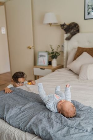 A baby lies on a gray comforter while a cat lounges on the headboard of the bed in a cozy bedroom setting.