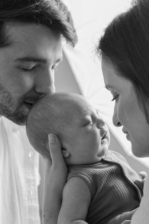 A tender black and white portrait of parents sharing an intimate moment with their sleeping newborn baby.