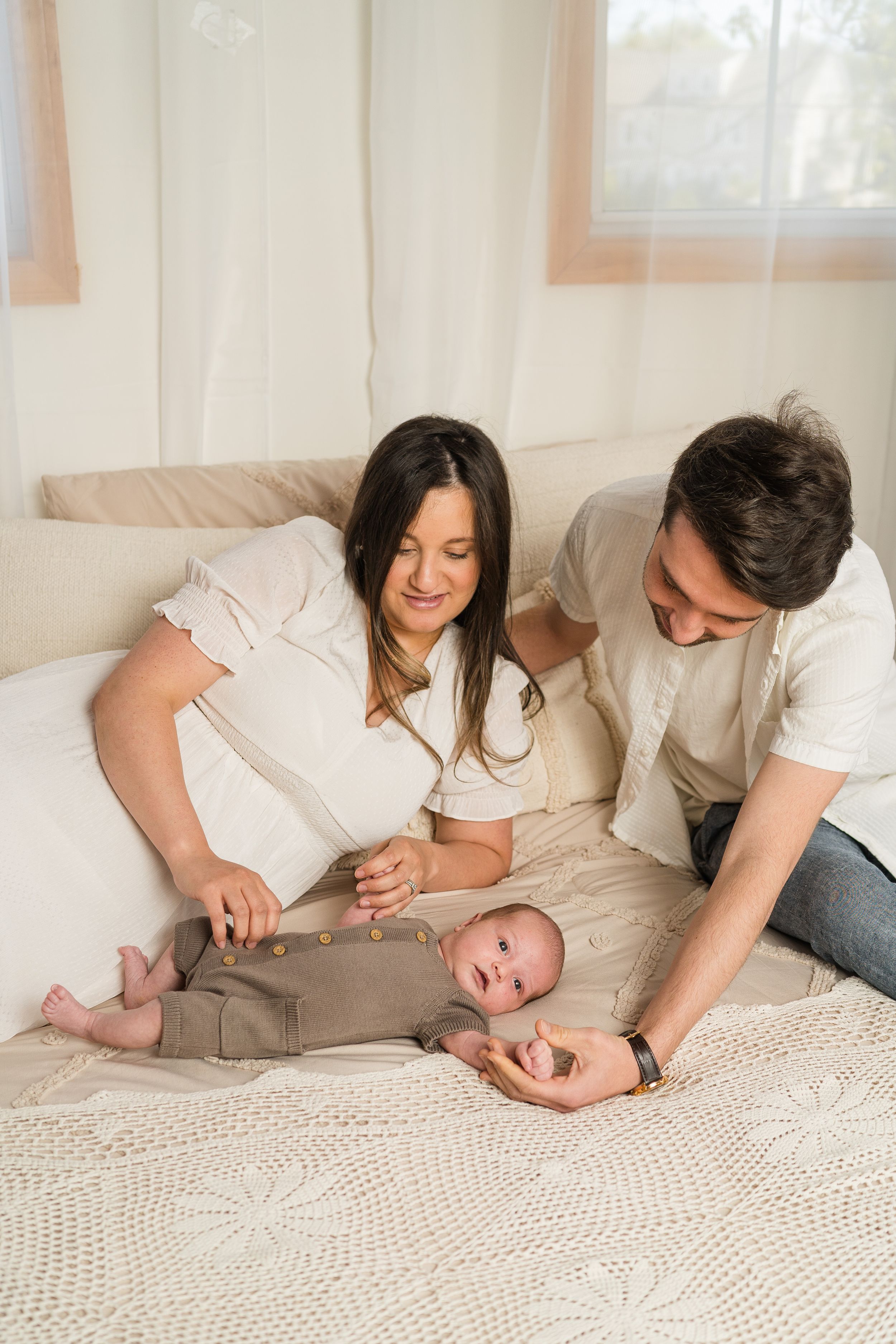 Parents playing and bonding with baby on white bedding in a bright, airy room.