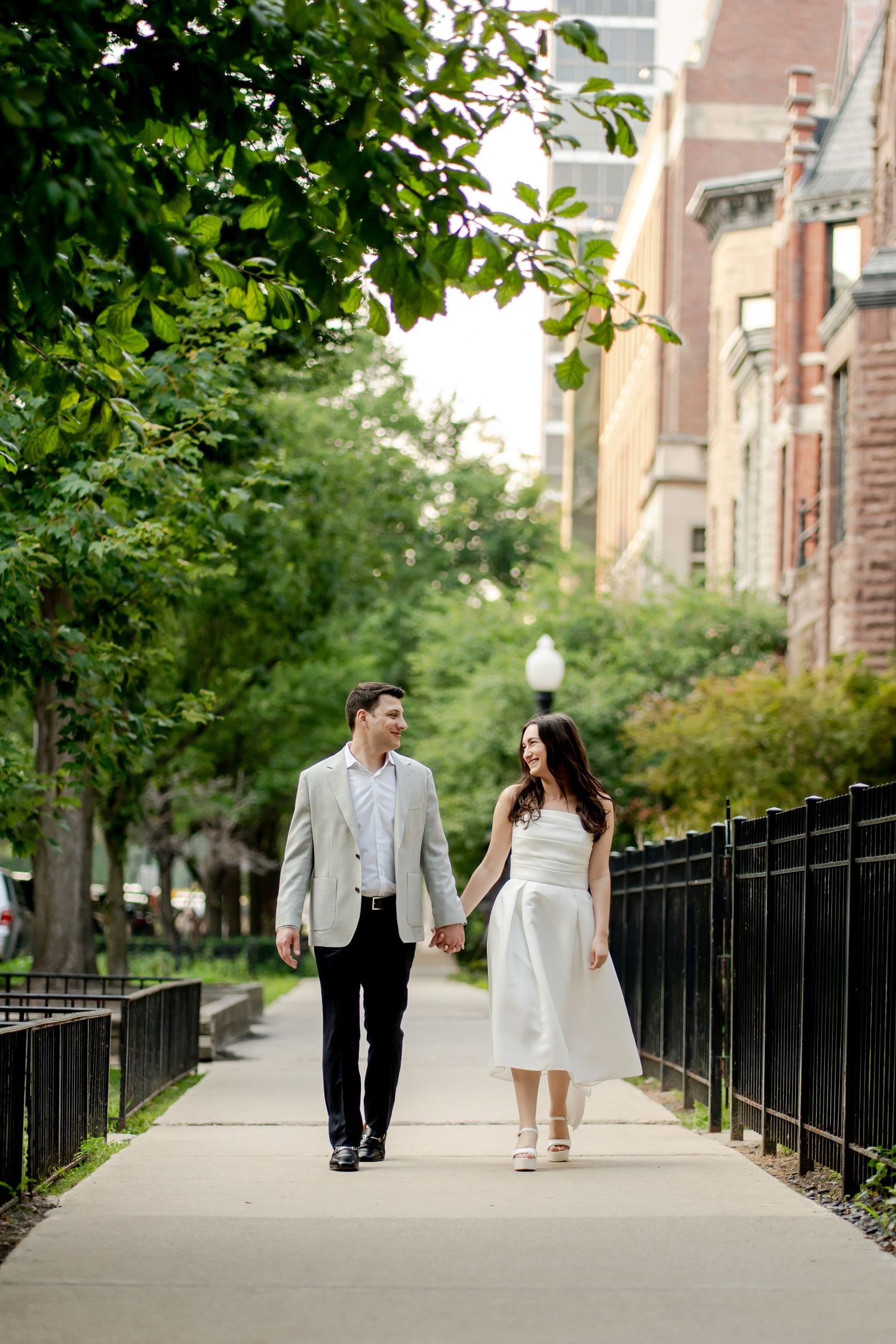 Smiling couple holding hands walking down a tree-lined city sidewalk past brick buildings, she in a white dress and he in a light blazer.