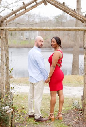 Couple poses playfully under rustic wooden arbor in outdoor engagement photo series.