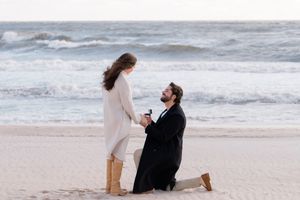 A proposal moment unfolds on a sandy beach with waves crashing in the background by Hamptons photographer.