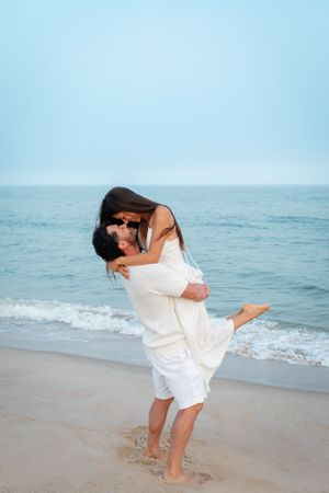A couple in white embraces and shares a romantic moment on a serene beach with ocean waves in the background.