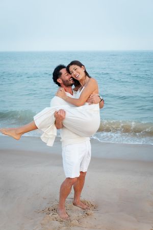 A romantic couple in white outfits embracing on the beach with ocean waves in the background.