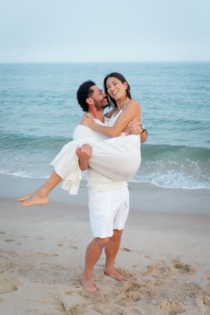 A romantic couple in white outfits embraces on the beach with ocean waves in the background.