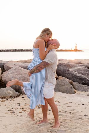 A couple in coordinated blue and white outfits embracing on a rocky beach during sunset by Hamptons photographer.