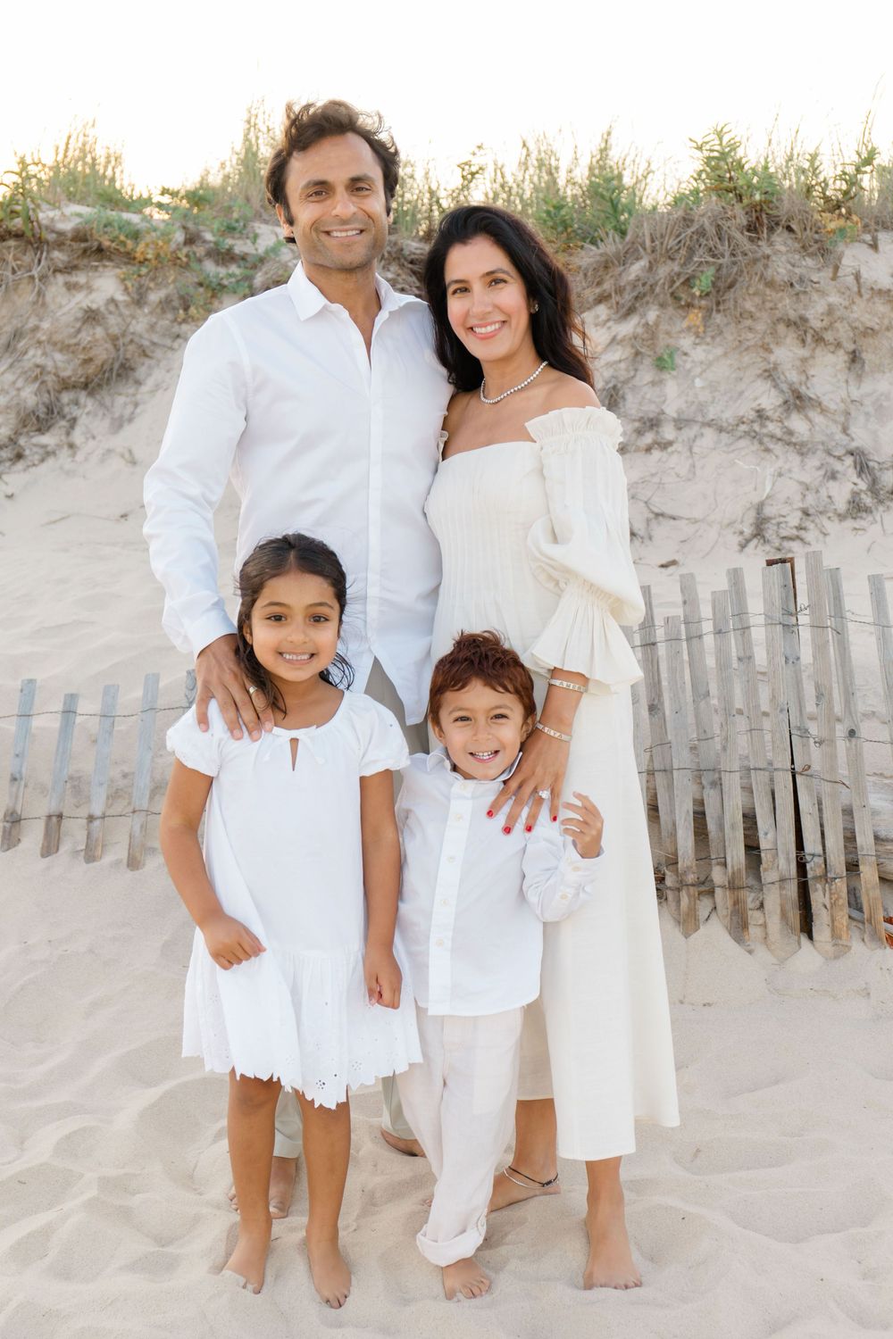 Family wearing all white outfits poses together on sandy beach dunes near wooden fence.