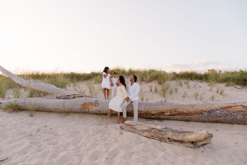 Family in white clothing poses together on driftwood log at beach during sunset.