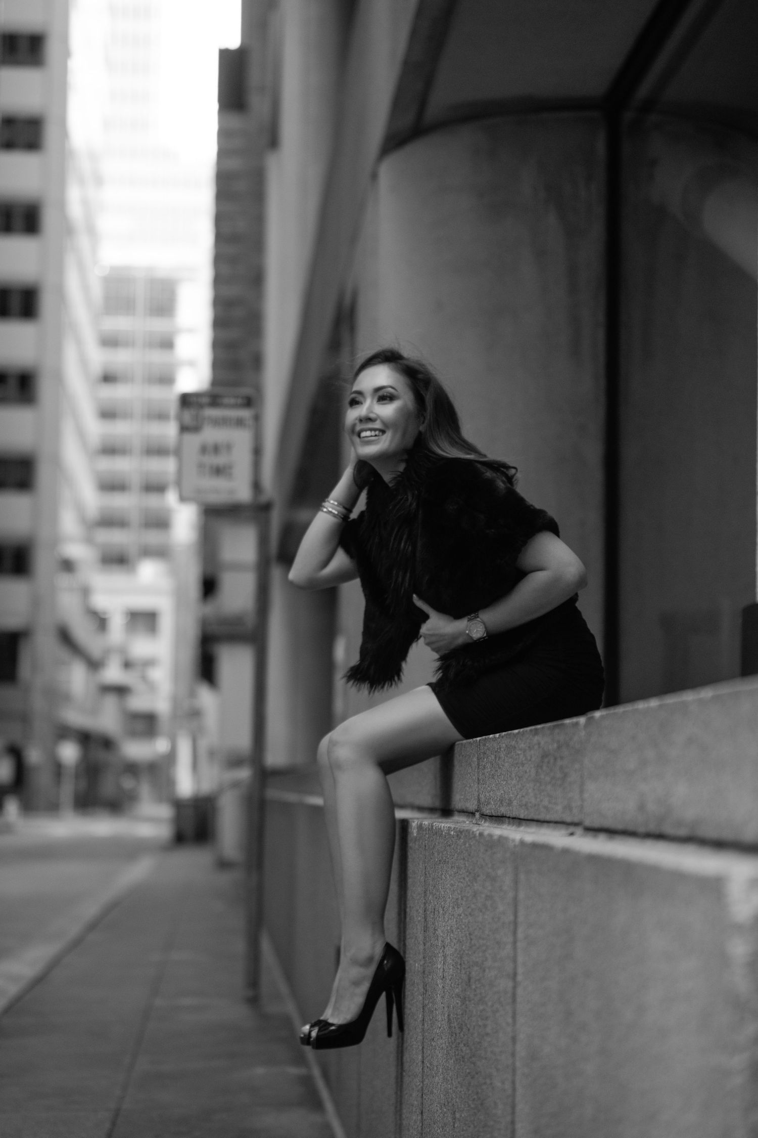 Black and white photo of a smiling woman in a black dress and heels sitting on a ledge in an urban city setting.