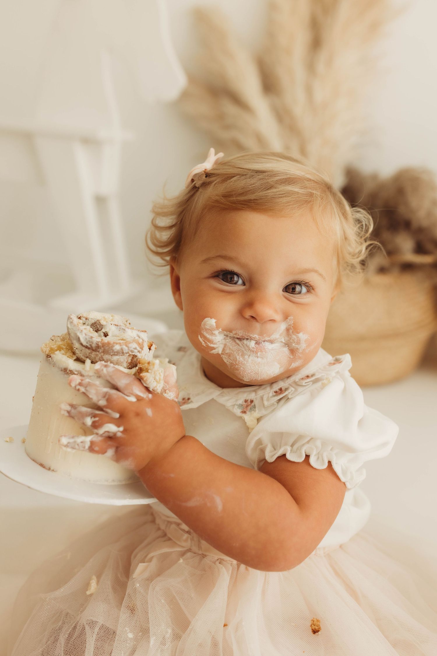 Baby in white lace dress reaching excitedly for birthday cake on white stand.