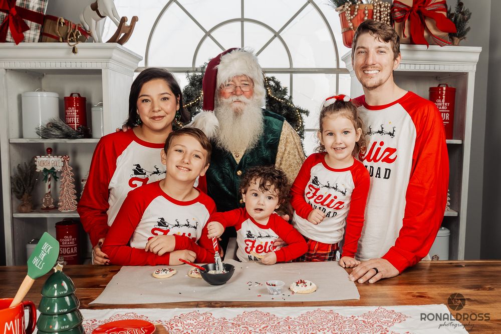 A group wearing matching red and white holiday shirts sits together on a couch in front of Christmas decorations. Santa Photos League City
