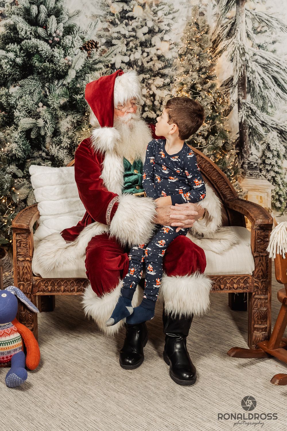 Festive holiday photo session with Santa Claus and a child on wooden bench surrounded by snowy Christmas trees. Santa Photos Webster