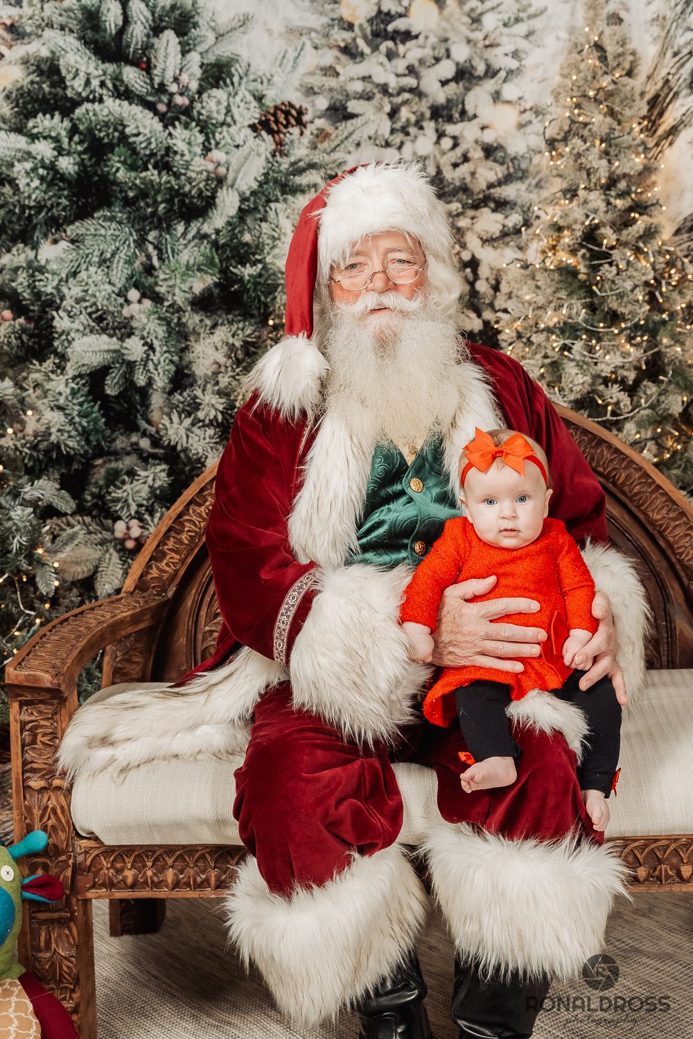 A baby in an orange outfit sits on a traditional Christmas scene with snowy evergreen trees in the background. Santa Photos Galveston