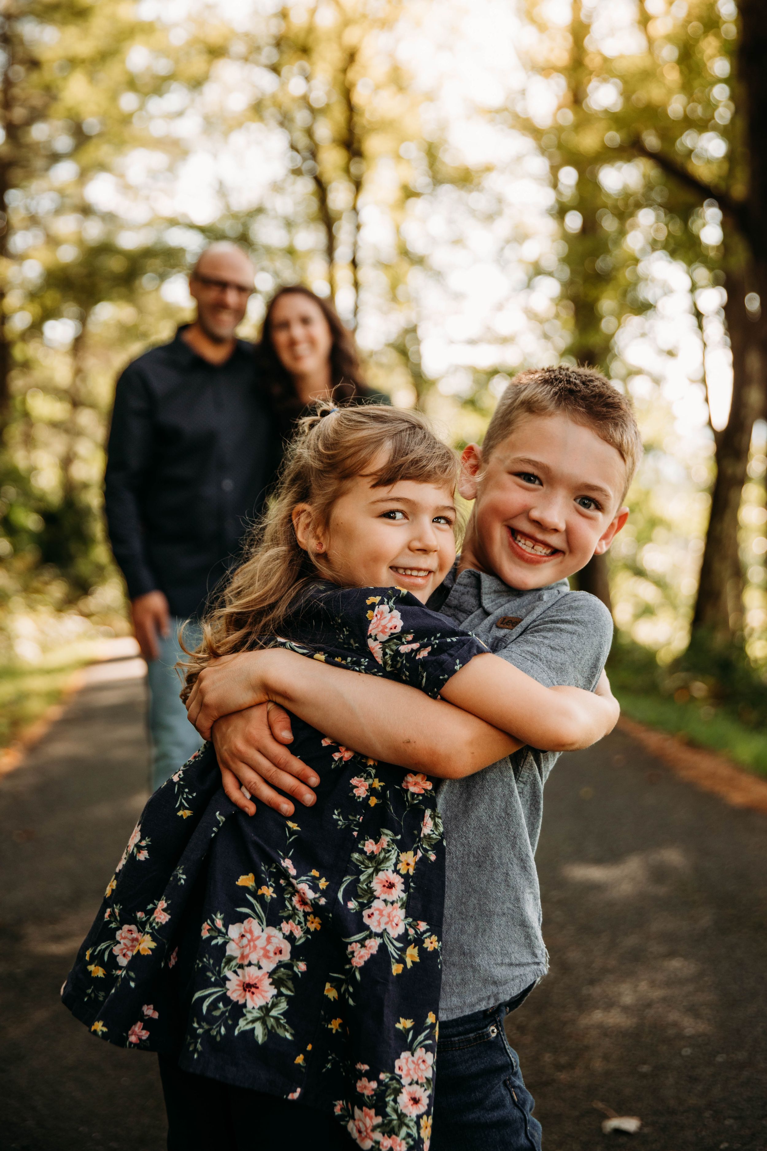 Two young siblings hugging and smiling on a forest path with parents standing behind them.