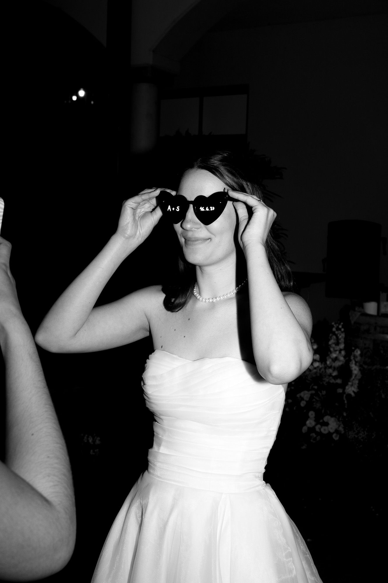 A bride in a white dress tries on heart-shaped sunglasses in black and white.