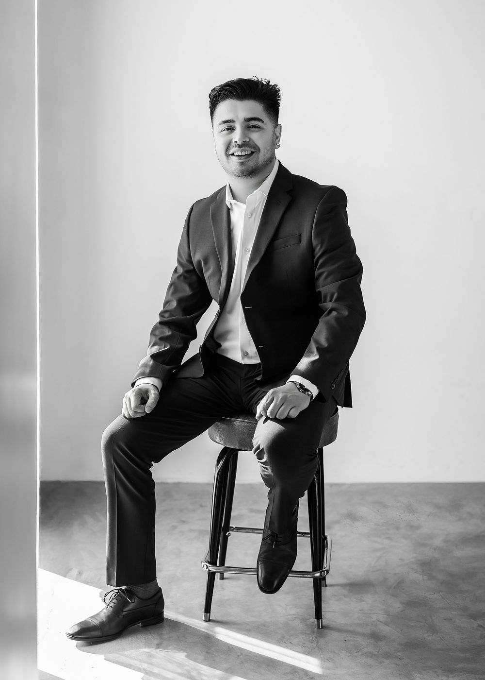 A smiling man in a black suit sits on a stool in a bright studio, posing confidently for a professional portrait.