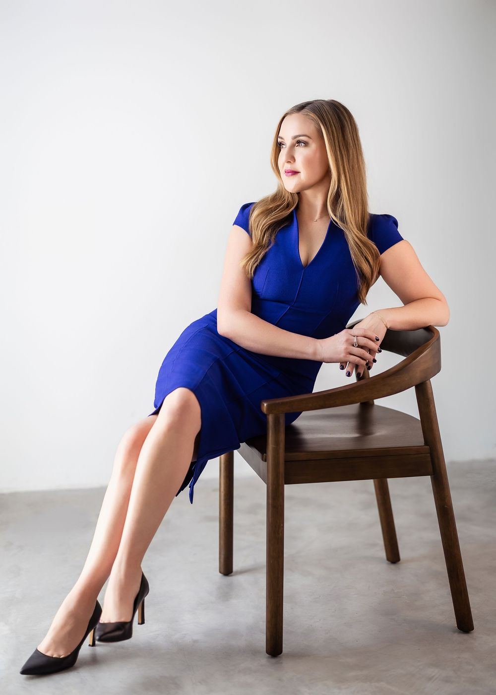 Woman in royal blue dress seated on wooden chair, posing confidently in a bright studio setting.
