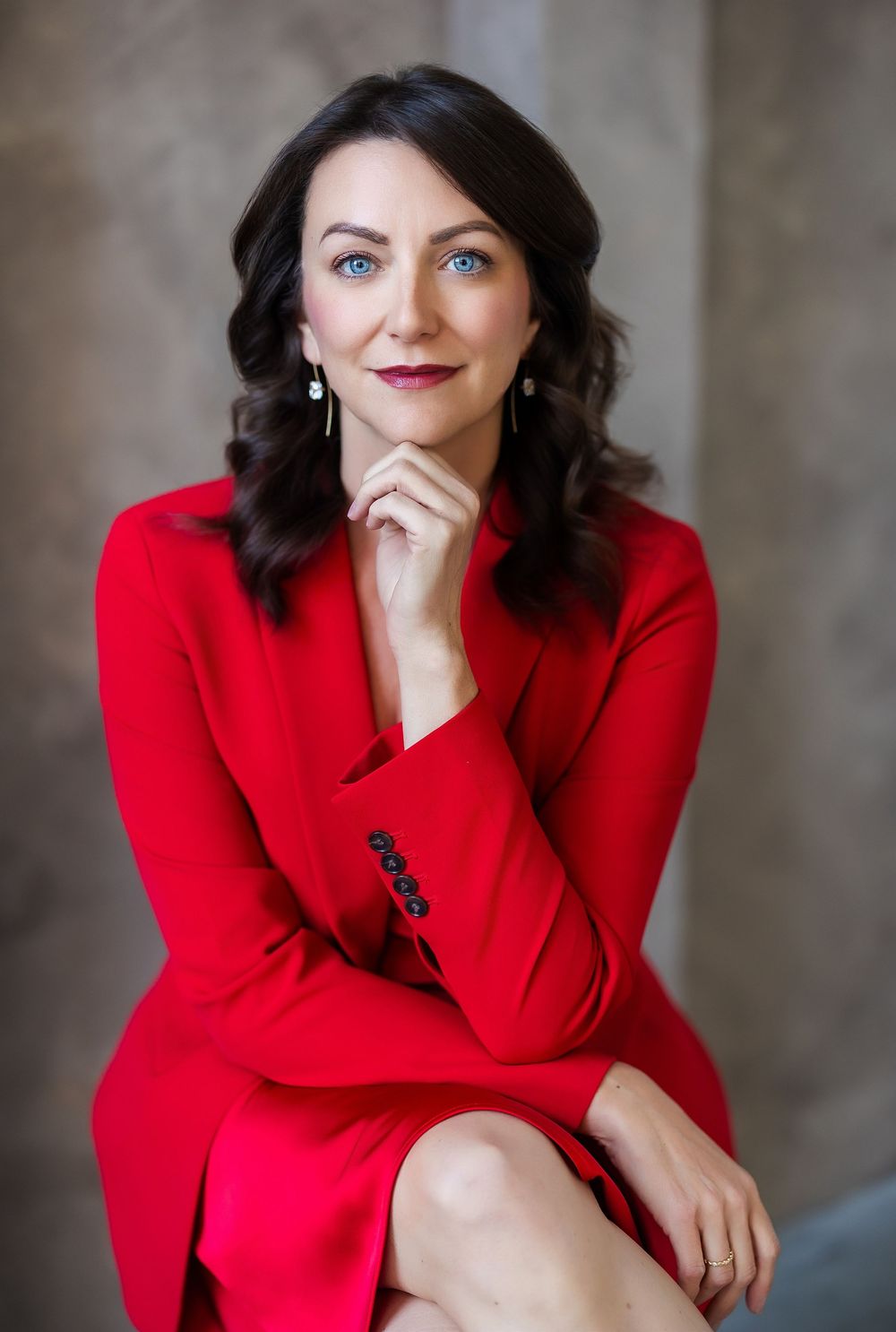 Professional woman in red blazer dress posing confidently with hand on chin, dark wavy hair and blue eyes.