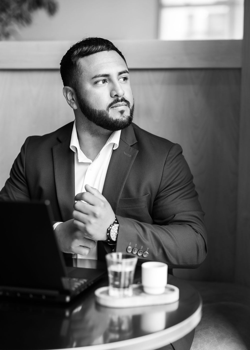 Professional in a dark suit jacket sits thoughtfully at a cafe table with a beverage in black and white.