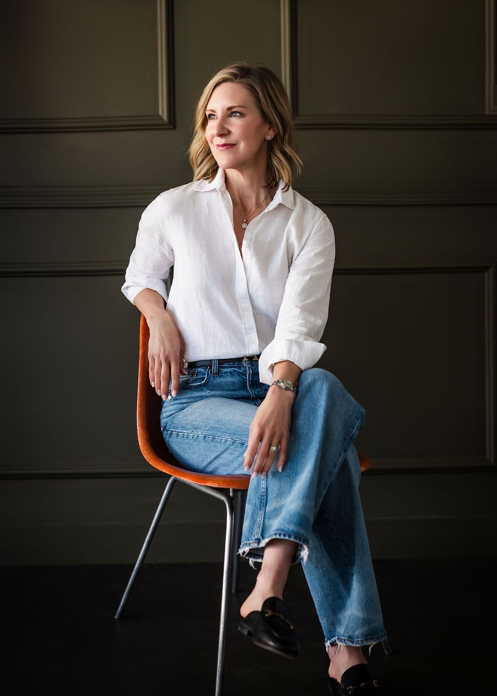 Woman in white shirt and jeans sitting on orange chair against dark green wall in professional portrait.