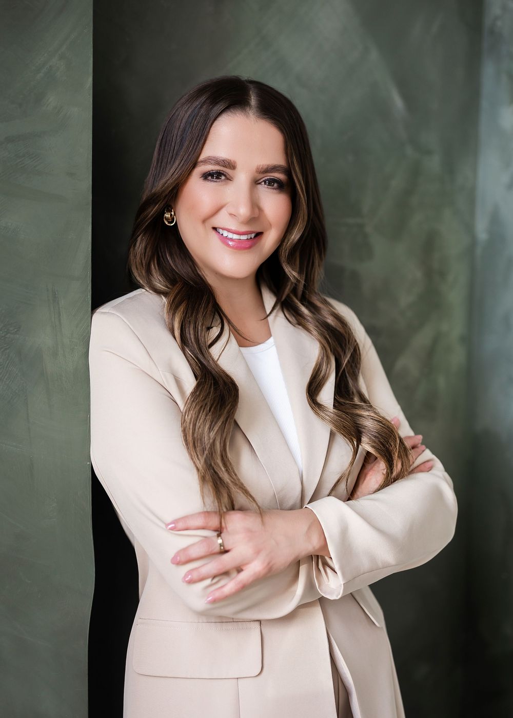 Professional woman in beige blazer with crossed arms, smiling confidently against a green backdrop.