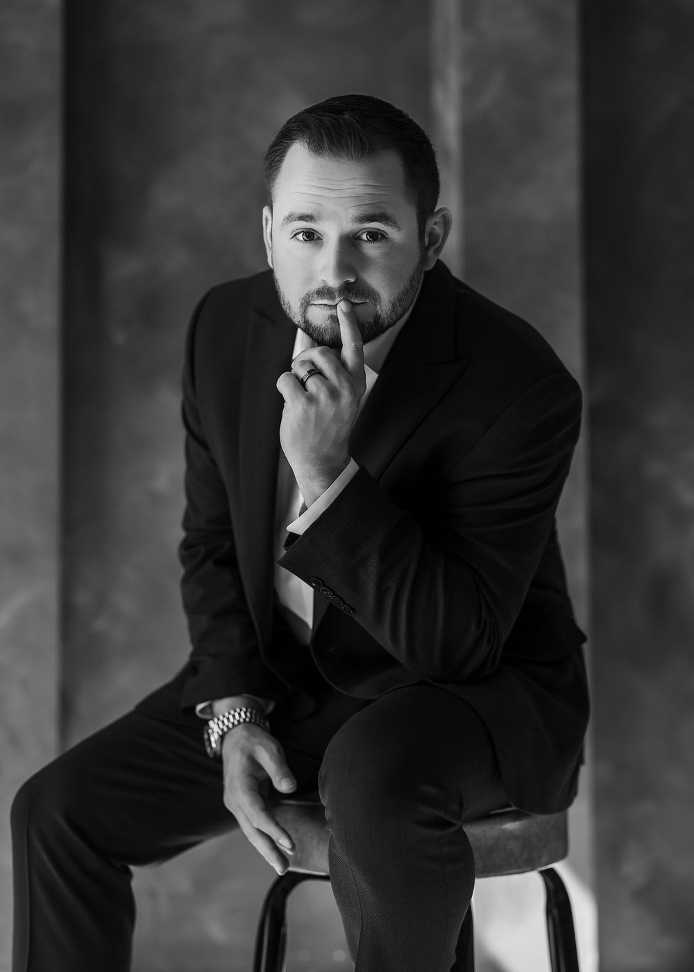 Black and white portrait of a man in a suit sitting on a stool, hand on chin, in a thoughtful pose.