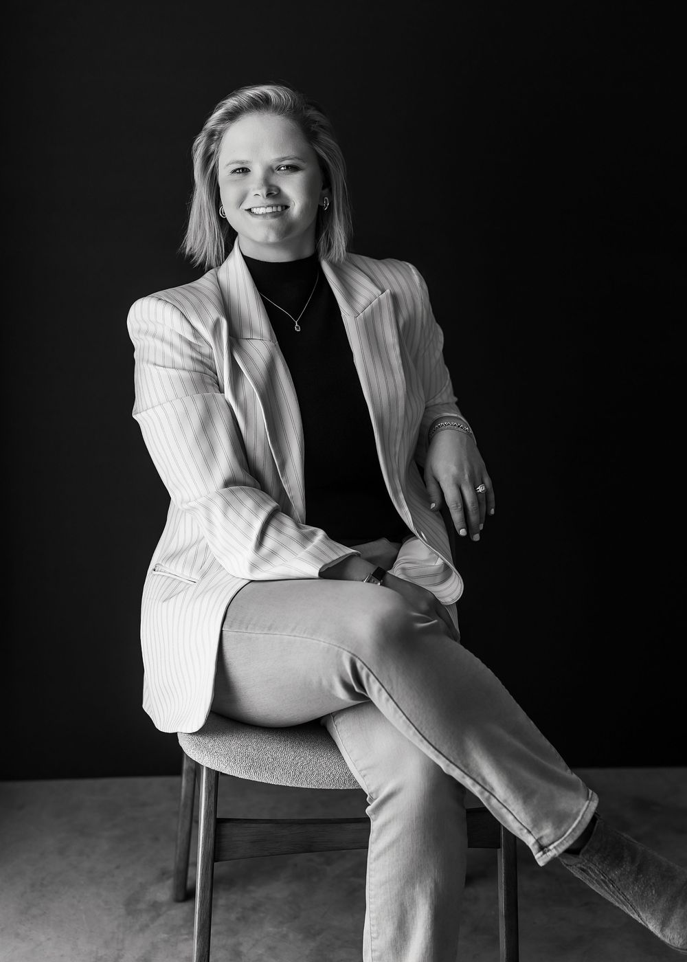 Black and white portrait of a smiling blonde woman in a blazer sitting confidently on a chair in a studio.