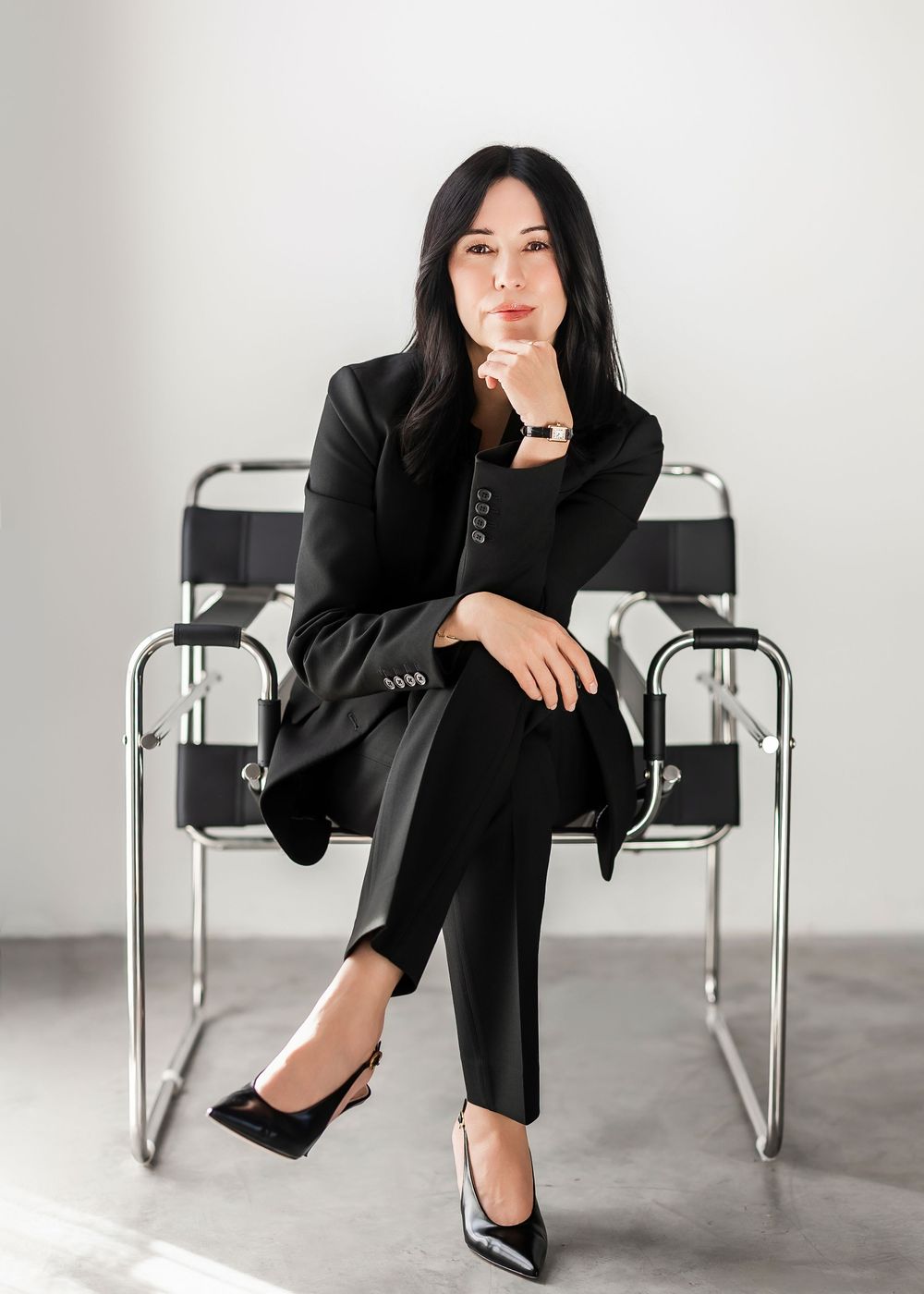 Woman in black suit sitting confidently on a chrome Wassily chair against a white background.